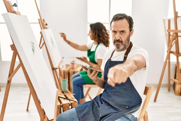 Hispanic middle age man and mature woman at art studio pointing with finger to the camera and to you, confident gesture looking serious