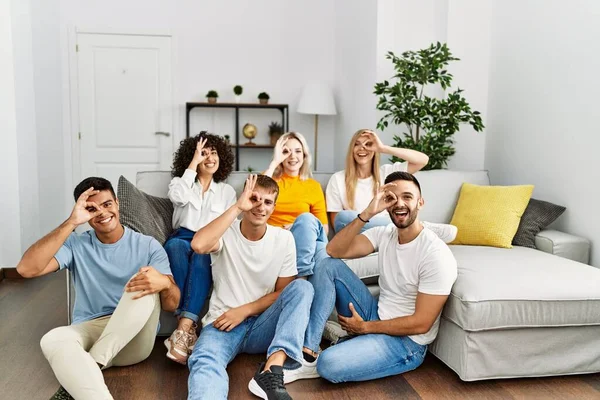 Group of people sitting on the sofa and floor at home smiling happy doing ok sign with hand on eye looking through fingers