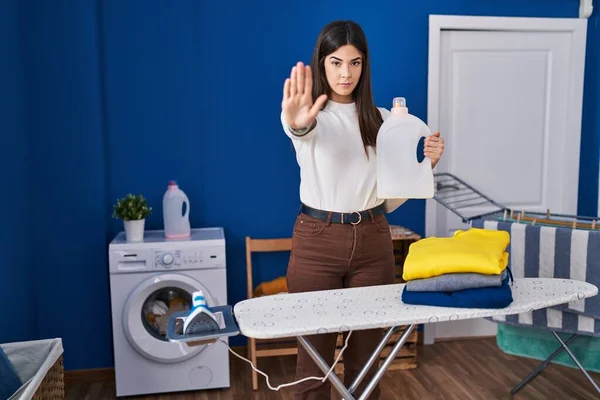 Young brunette woman with folded laundry after ironing with open hand doing stop sign with serious and confident expression, defense gesture