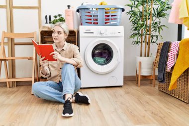 Çamaşır odasında çamaşır makinesi bekleyen, kitap okuyan beyaz bir kadın.