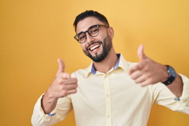 İş kıyafetleri ve gözlük takan İspanyol genç adam mutlu ve komik yüzüyle kamerayı işaret ediyor. iyi enerji ve titreşimler. 