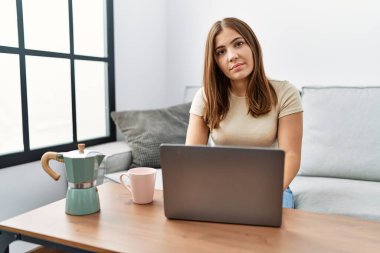 Genç esmer kadın evde dizüstü bilgisayar kullanıyor. Bir fincan kahve içiyor. Yüzünde ciddi bir ifade var. Basit ve doğal bir şekilde kameraya bakıyorsun.. 
