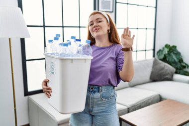 Genç kızıl saçlı kadın elinde plastik şişelerle çöp sepetini tutarak selam veriyor mutlu ve güler yüzlü, dostça bir jest yapıyor. 