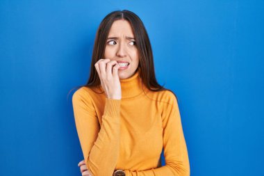 Mavi arka planda duran esmer kadın gergin ve gergin görünüyor. Ağzı tırnak yiyen ellerle. anksiyete. 