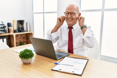 Senior man working at the office using computer laptop smiling pulling ears with fingers, funny gesture. audition problem 