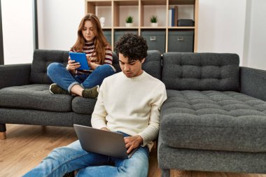 Ciddi ifadeleri olan genç bir çift evdeki koltukta dizüstü bilgisayar ve dokunmatik ped kullanıyor..