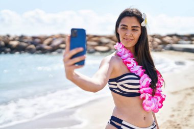 Bikini giyen genç İspanyol kadın deniz kenarında akıllı telefonla selfie çekiyor.