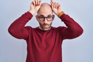 Beyaz arka plan üzerinde duran sakallı genç kel bir adam gözlük takıyor ve boynuzları gibi parmağıyla komik hareketler yapıyor. 