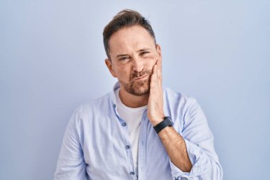 Orta yaşlı, mavi arka planda duran beyaz bir adam diş ağrısı ya da diş ağrısı yüzünden acı dolu bir ifadeyle ağıza dokunuyor. dişçi 