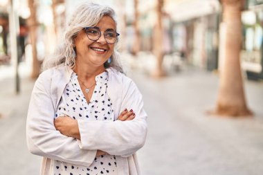 Orta yaşlı kadın gülümsüyor. Kollarını kavuşturup caddede duruyor.