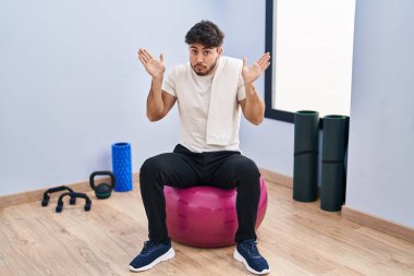 Hispanic man with beard sitting on pilate balls at yoga room clueless and confused expression with arms and hands raised. doubt concept. 