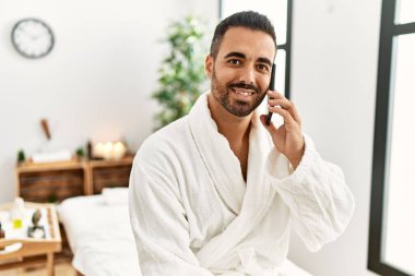 Young hispanic man wearing bathrobe talking on the smartphone at beauty center