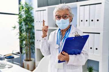 Gri saçlı, doktor üniforması giyen, güvenlik maskesi takan, bir fikir veya soruyla gülümseyen, mutlu yüzlü, yaşlı bir kadın. 