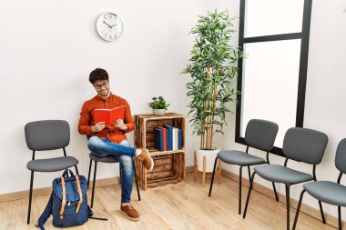 Young hispanic man smiling confident reading book at waiting room
