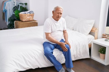 Senior grey-haired man suffering for knee injury sitting on bed at bedroom