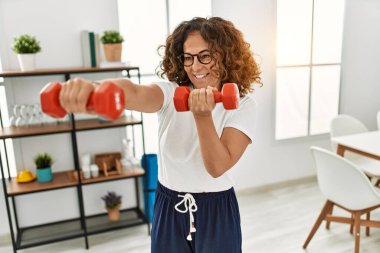 Orta yaşlı İspanyol kadın gülümsüyor. Eğitimine özgüveni tam. Evde dambıl kullanıyor.