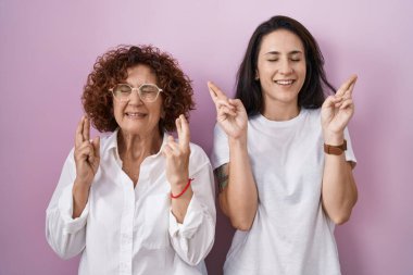 İspanyol anne ve kızı pembe arka planda gelişigüzel beyaz tişörtler giyip umut ve gözlerin kapalı gülümseyerek el kol hareketi yapıyorlar. Şans ve batıl inançlar. 