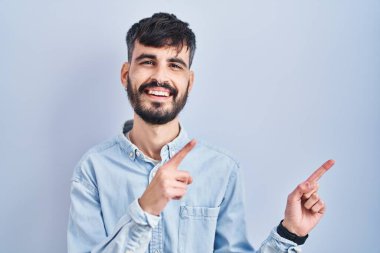Mavi arka planda duran, gülümseyen ve kameraya bakan, iki eli ve parmağı yanda olan Latin kökenli genç bir adam.. 