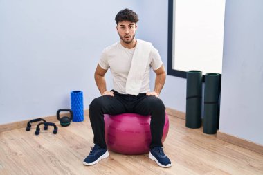 Hispanic man with beard sitting on pilate balls at yoga room afraid and shocked with surprise expression, fear and excited face. 