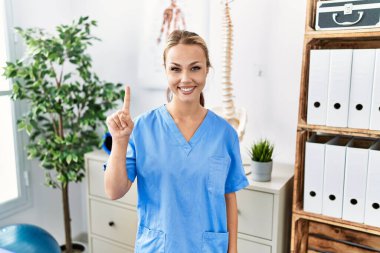 Ağrı kesici kliniğinde çalışan beyaz bir kadın bir numaralı parmağı gösterip işaret ederken kendinden emin ve mutlu bir şekilde gülümsüyor.. 