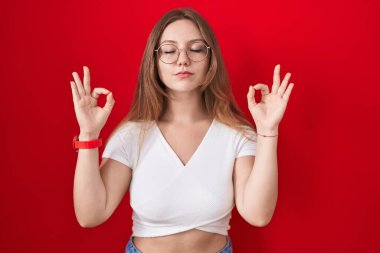 Kafkasyalı genç bir kadın kırmızı arka planda rahatlamış ve parmaklarıyla meditasyon hareketi yaparken gözleri kapalı gülümsüyor. yoga konsepti. 