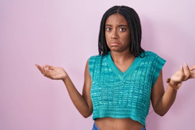 Saçları pembe arka planda duran, elleri ve kolları havada, kafası karışmış genç bir Afrikalı Amerikalı. şüphe kavramı. 