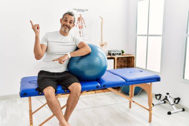 Orta yaşlı İspanyol bir adam ağrı kesici kliniğinde elinde kocaman bir gülümsemeyle pilates topu tutuyor, kameraya el ile işaret ediyor.. 