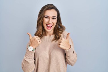 Young woman standing over isolated background success sign doing positive gesture with hand, thumbs up smiling and happy. cheerful expression and winner gesture. 