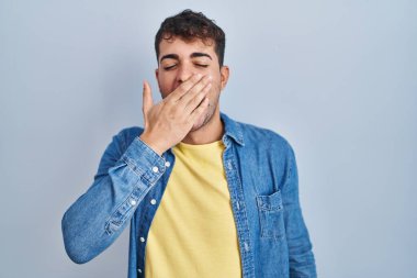 Mavi arka planda dikilen İspanyol genç adam ağzıyla ağzını kapatmaktan sıkılmış. Huzursuz ve uykulu.. 
