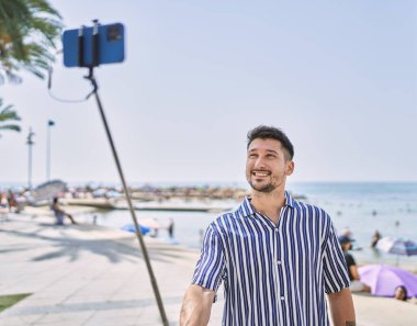 Genç yakışıklı adam deniz kenarında selfie çubuğu kullanarak fotoğraf çekiyor.