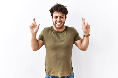 Hispanic man standing over isolated white background gesturing finger crossed smiling with hope and eyes closed. luck and superstitious concept. 