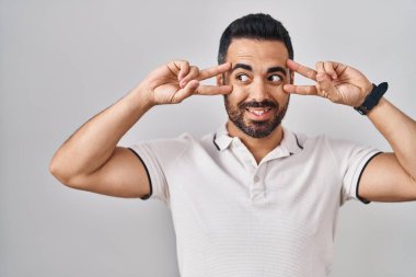 Sakallı, beyaz arka planda günlük giysiler giyen, yüzünde parmaklarıyla barış sembolü çizen, neşeli gülümseyen, zaferi gösteren genç İspanyol bir adam. 