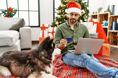 Genç İspanyol adam dizüstü bilgisayar ve kredi kartı kullanıyor evdeki yılbaşı ağacında köpekle birlikte yerde oturuyor.