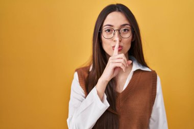 Sarı arka planda duran genç esmer kadın gözlüklü ve sessiz olmak istiyor. sessizlik ve gizli konsept. 