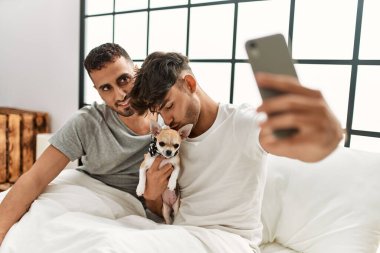 İki İspanyol erkek yatak odasında chihuahua ile yatakta otururken akıllı telefonun yanında selfie çekiyorlar.