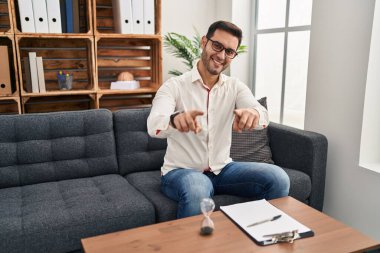 Danışma ofisinde çalışan sakallı genç İspanyol adam seni ve kamerayı işaret ediyor, gülümsüyor, pozitif ve neşeli. 