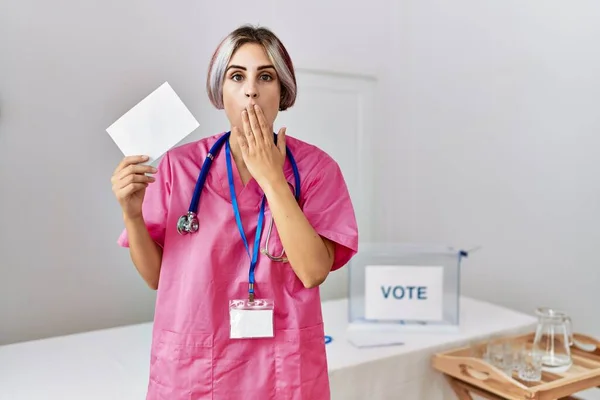 Young nurse woman at political campaign election holding envelope covering mouth with hand, shocked and afraid for mistake. surprised expression