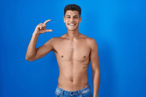 Young hispanic man standing shirtless over blue background smiling and confident gesturing with hand doing small size sign with fingers looking and the camera. measure concept.
