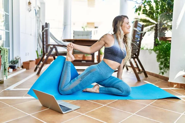 Beautiful caucasian woman doing exercise and stretching at the terrace at her home. Doing asana and wellbeing work out on yoga mat looking at traning tutorial on laptop.