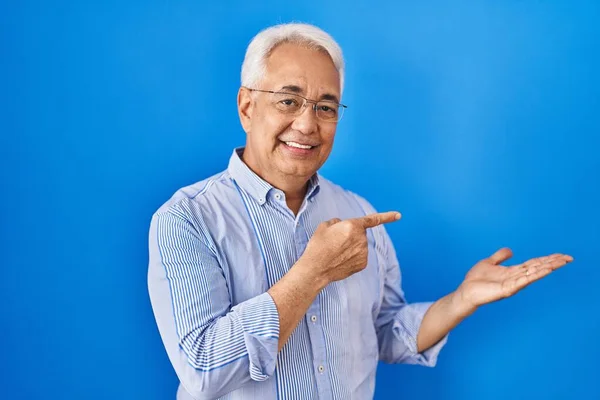 Hispanic senior man wearing glasses amazed and smiling to the camera while presenting with hand and pointing with finger.