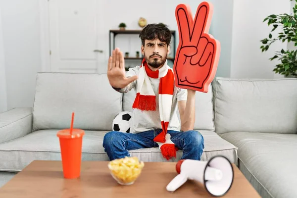 Hispanic man with beard supporting football team at home sitting on the sofa with open hand doing stop sign with serious and confident expression, defense gesture