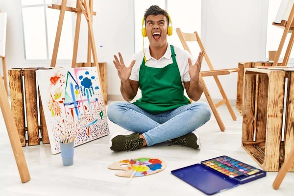 Young hispanic man at art studio crazy and mad shouting and yelling with aggressive expression and arms raised. frustration concept.