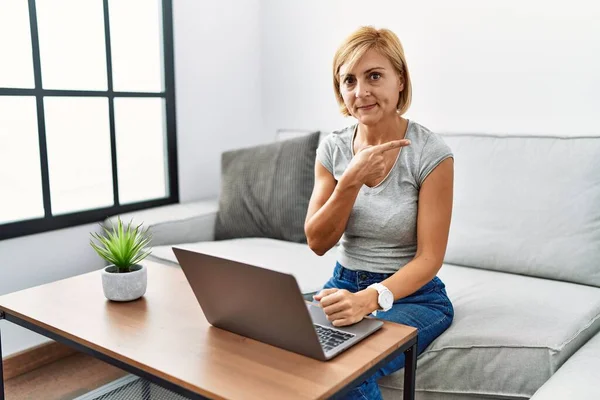 Middle age blonde woman using laptop at home pointing with hand finger to the side showing advertisement, serious and calm face