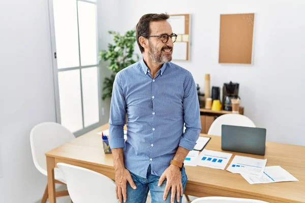 Middle age hispanic man with beard wearing business clothes at the office looking to side, relax profile pose with natural face with confident smile.