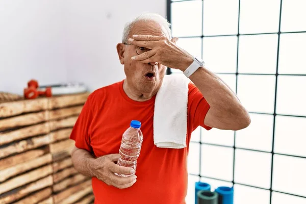 Senior man wearing sportswear and towel at the gym peeking in shock covering face and eyes with hand, looking through fingers with embarrassed expression.