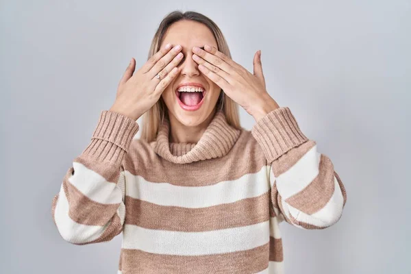 Young blonde woman wearing turtleneck sweater over isolated background covering eyes with hands smiling cheerful and funny. blind concept.