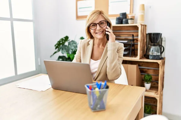 Middle age blonde woman smiling confident talking on the smartphone at office