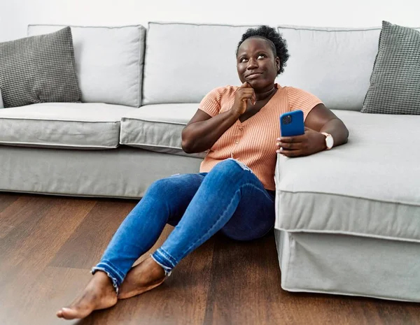 年轻的非洲女人用智能手机坐在家里的地板上手托着下巴思考着问题 沉思着 脸上挂着沉思的笑容 怀疑概念 — 图库照片