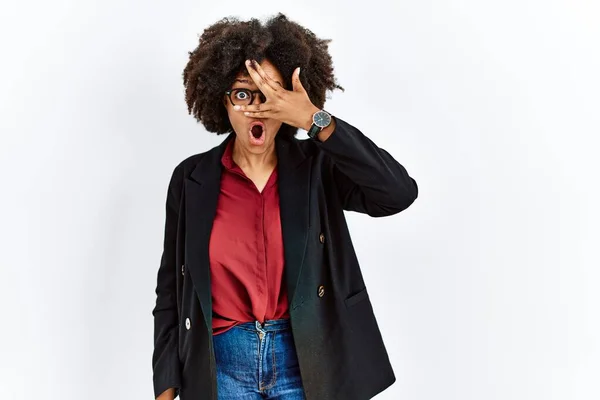 African american woman with afro hair wearing business jacket and glasses peeking in shock covering face and eyes with hand, looking through fingers with embarrassed expression.