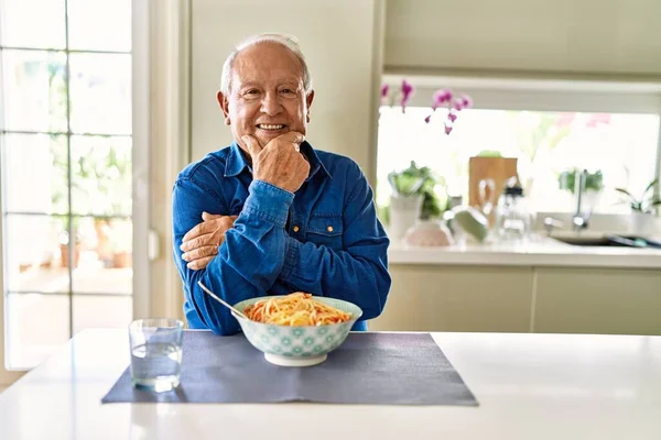 Senior man with grey hair eating pasta spaghetti at home looking confident at the camera smiling with crossed arms and hand raised on chin. thinking positive.